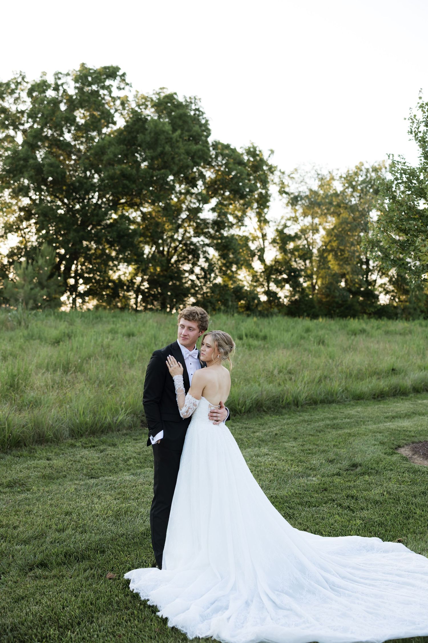 Bride and groom embracing at sunset in lush green field, bride wearing long white gown with train.