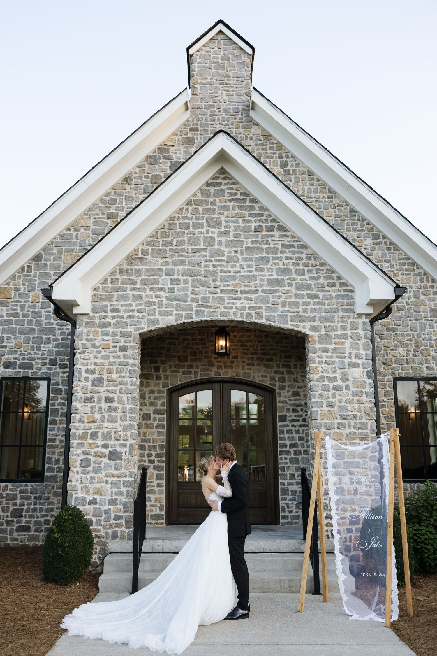 Bride and groom share romantic kiss outside rustic stone chapel with wedding sign display at dusk.