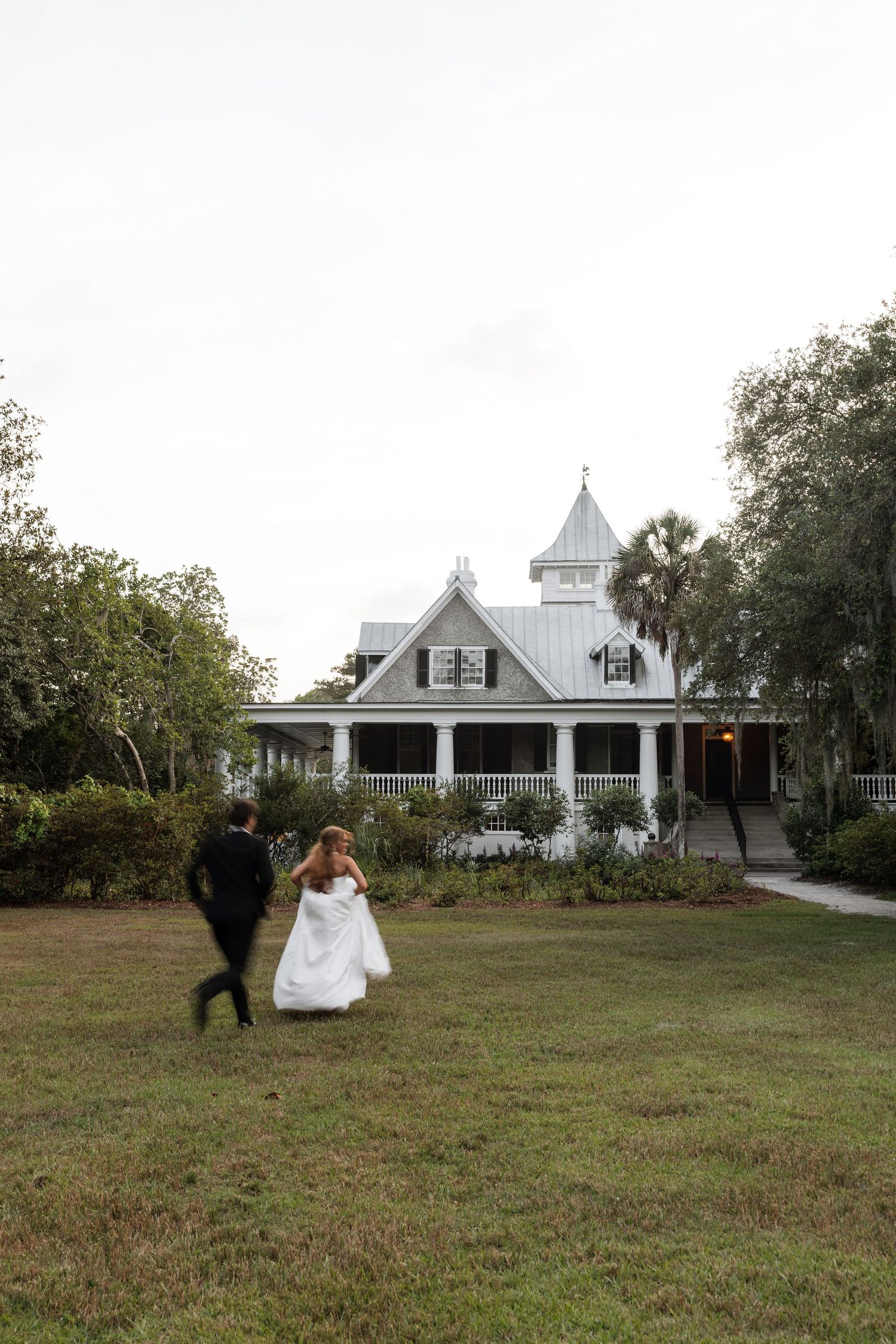Bride and groom running across green lawn toward charming white Victorian wedding venue surrounded by trees.