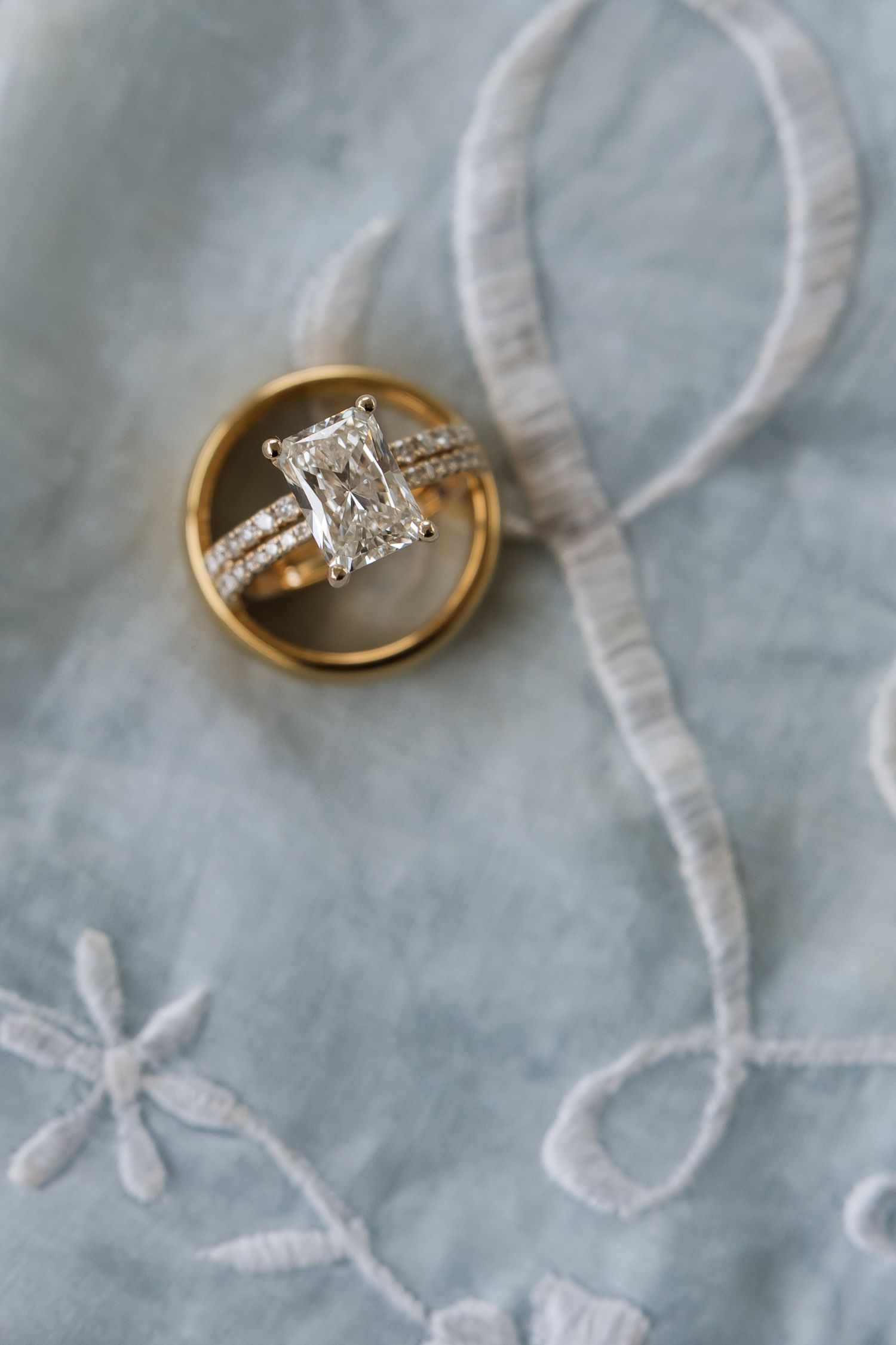 Gold diamond engagement ring with radiant cut stone and wedding band on embroidered blue fabric.