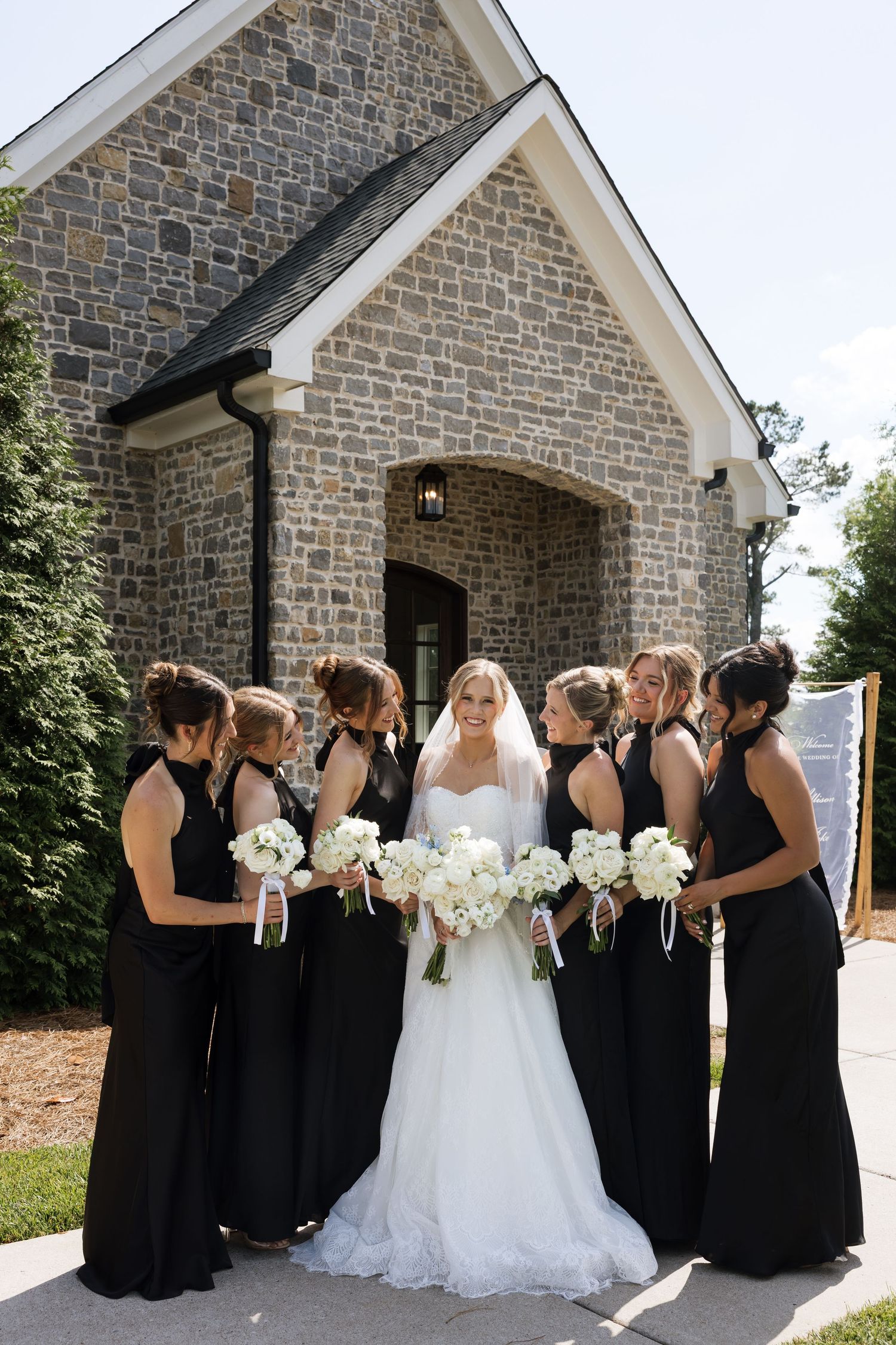 Bride in white gown with bridesmaids in black dresses holding white bouquets outside stone chapel.