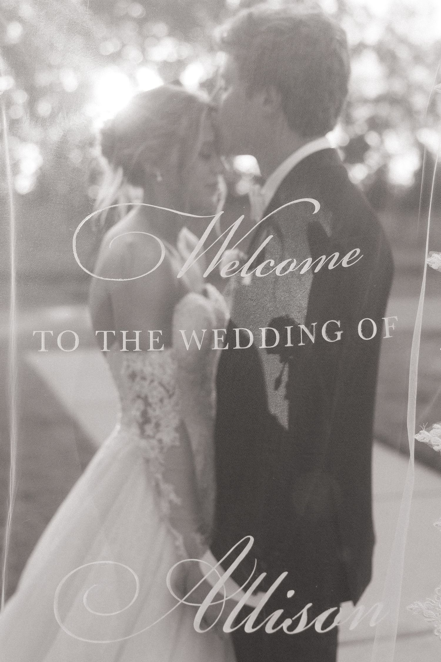 Black and white wedding welcome sign reading Welcome to the Wedding of Allison with couple kissing behind.