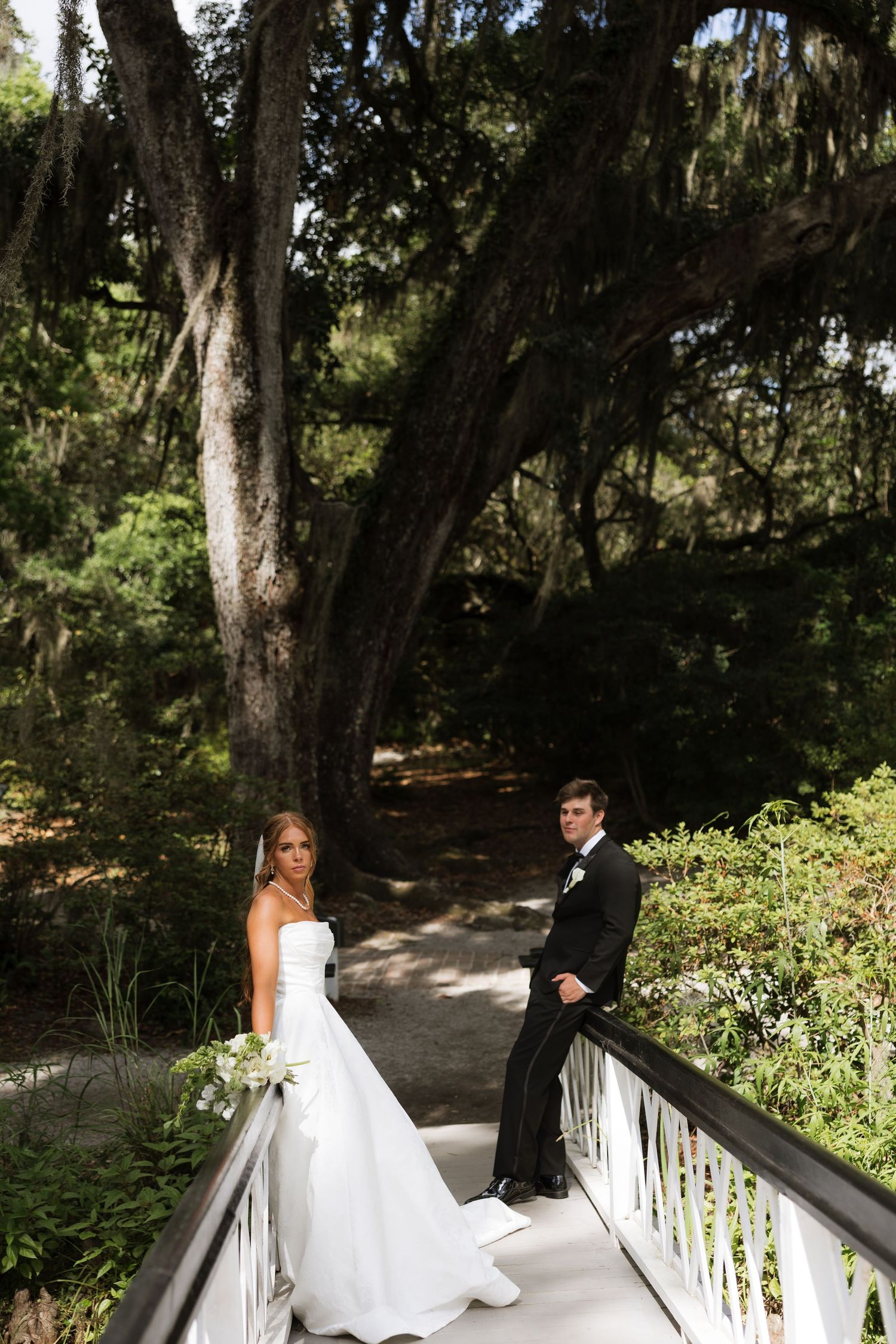 Bride and groom pose on a white bridge surrounded by lush trees with Spanish moss at an outdoor wedding.