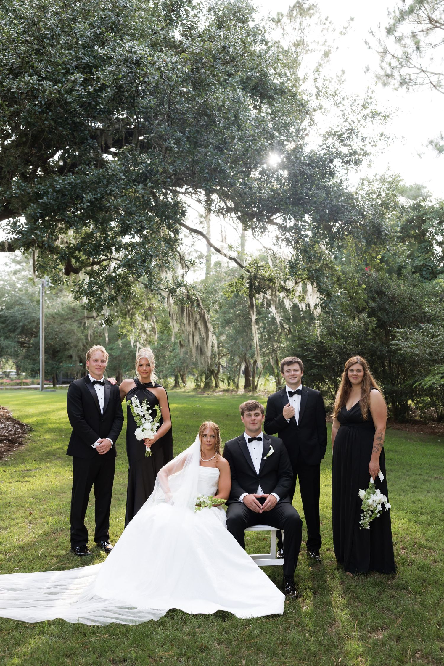 Wedding party in black formal wear poses with bride and groom under a large oak tree.