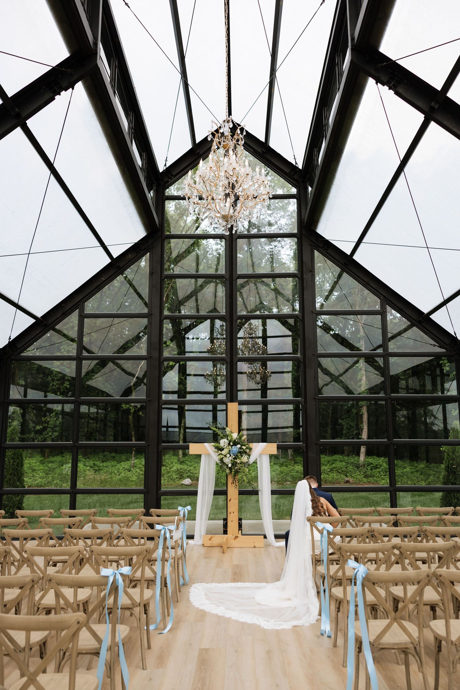 Elegant greenhouse wedding venue with glass ceiling, chandelier, wooden chairs with blue ribbons, and floral altar.