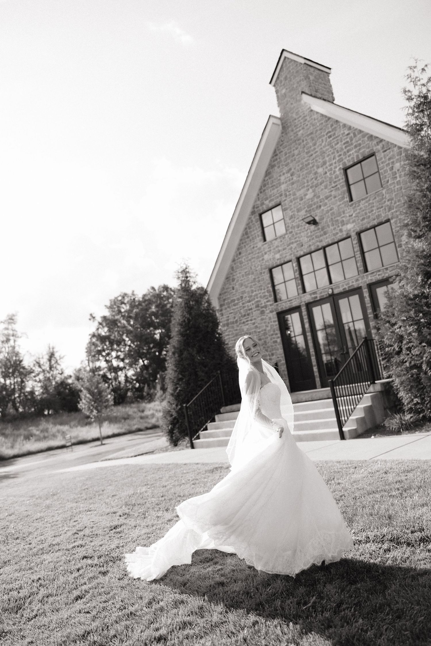 Bride in flowing white wedding gown and veil posing outside elegant brick venue in black and white.
