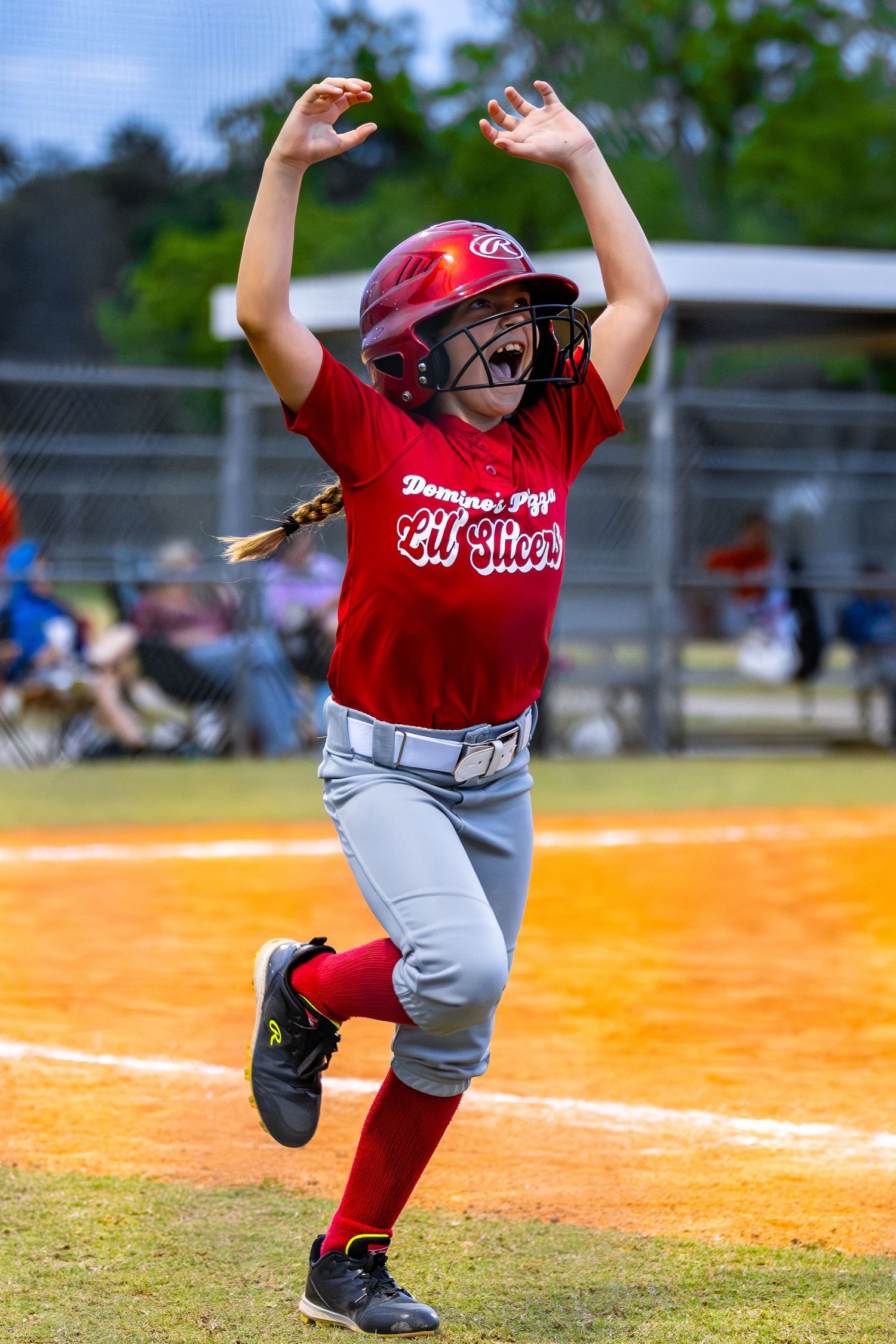 Young softball player in red uniform celebrating with arms raised while running bases during a youth league game.