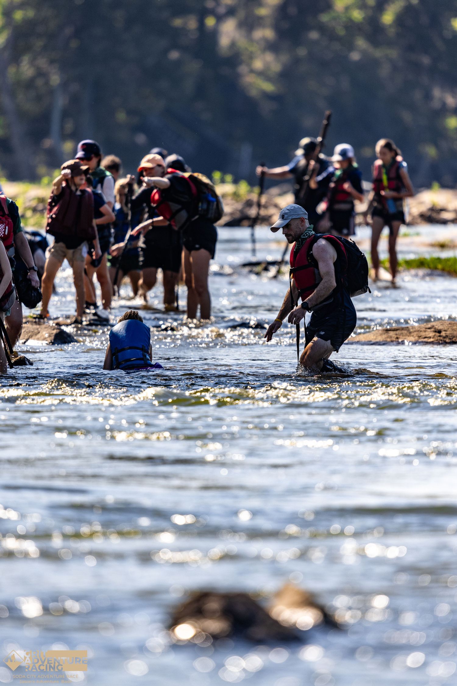 Large group of backpackers crossing a shallow river during an adventure race in a sunny wilderness setting.