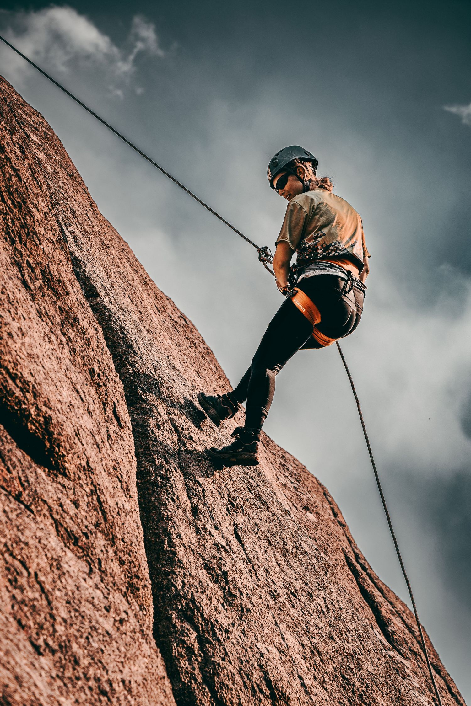 Rock climber rappelling down a steep granite cliff face with safety harness and helmet against cloudy sky.