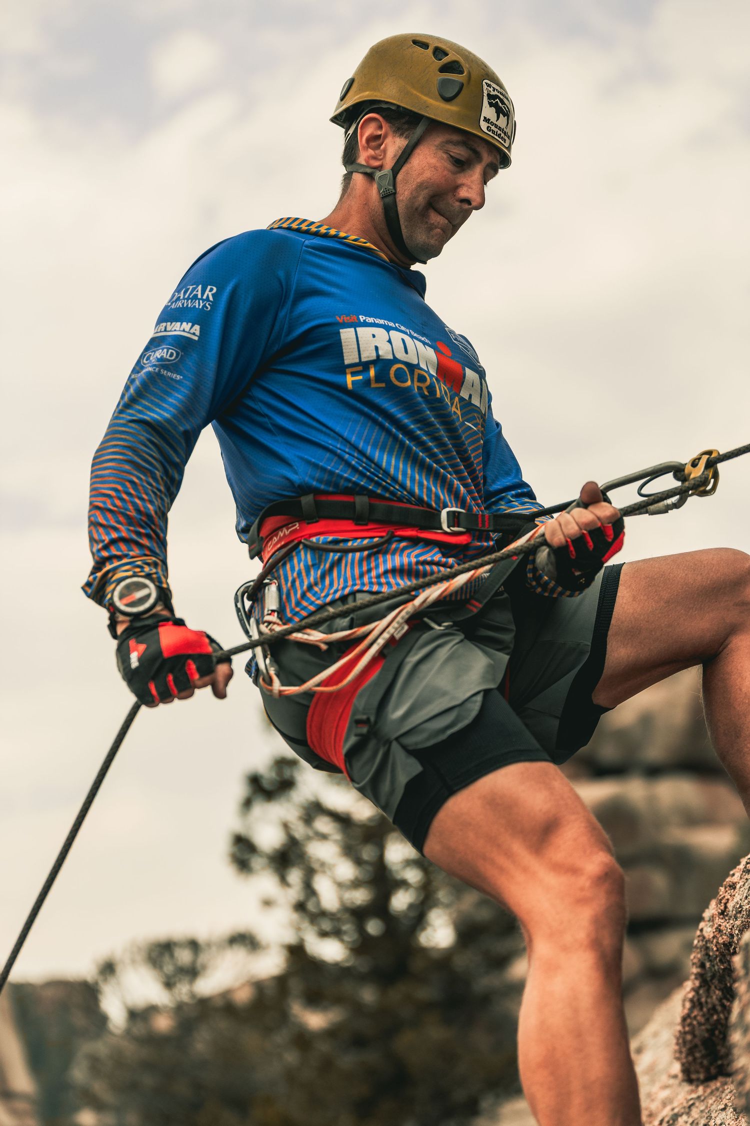 Male rock climber in blue Ironman shirt and helmet managing rappel rope on rocky cliff edge.