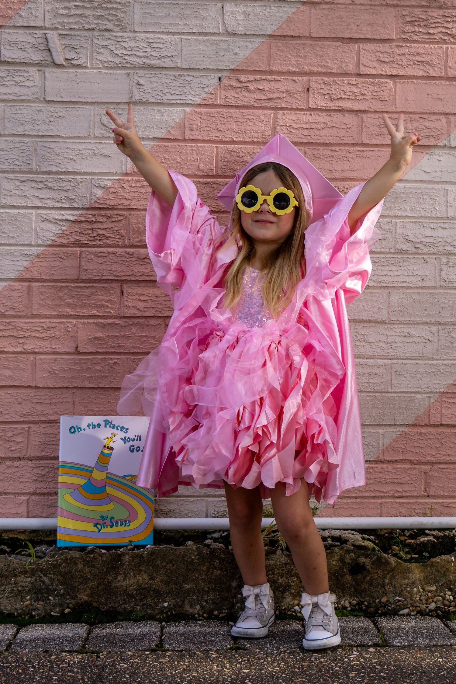Joyful young girl in pink graduation gown and cap with fun yellow sunglasses holding Dr. Seuss book.