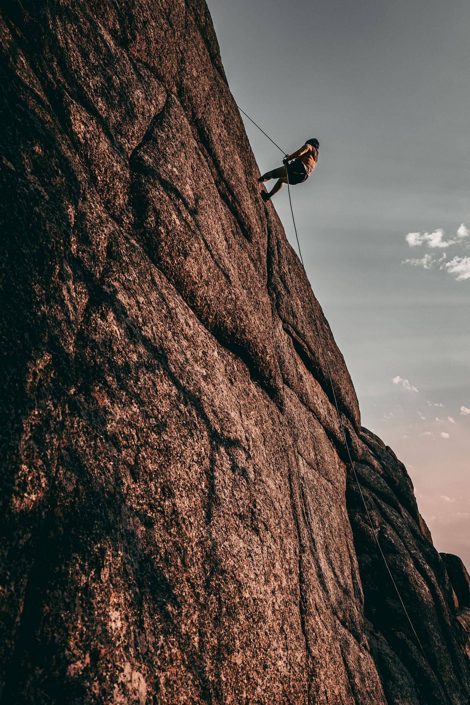 Solo climber rappelling down massive granite rock face at dusk with rope and harness.