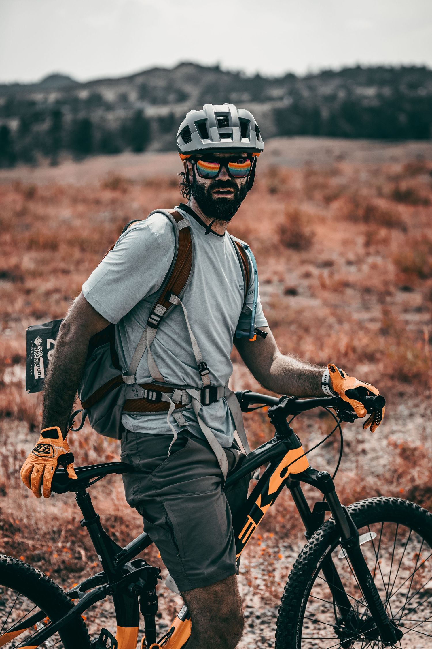 Bearded male mountain biker in helmet and orange gloves posing with bike in an autumn landscape.
