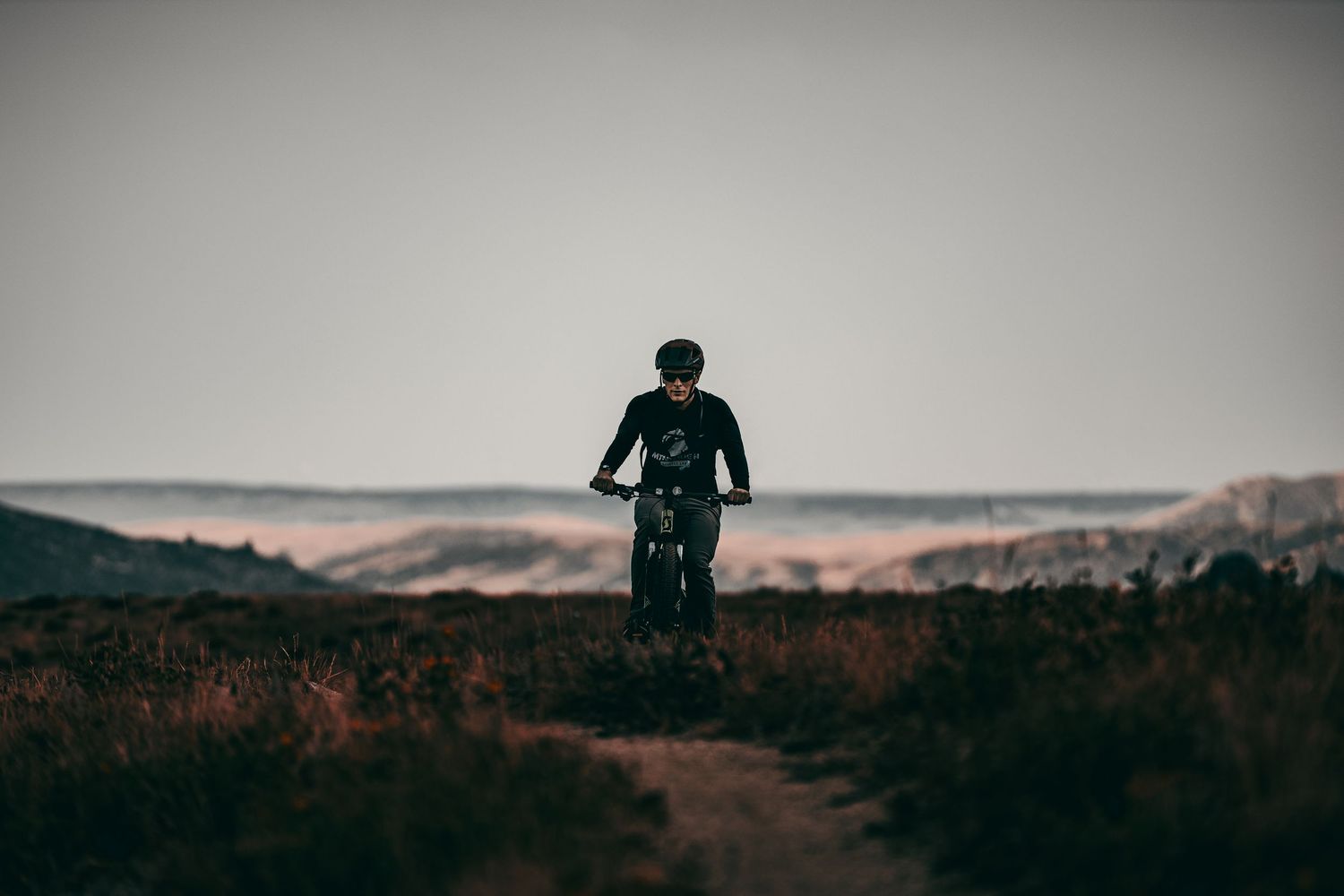 Lone mountain biker silhouetted against a moody sky on a rugged dirt trail in an open landscape.