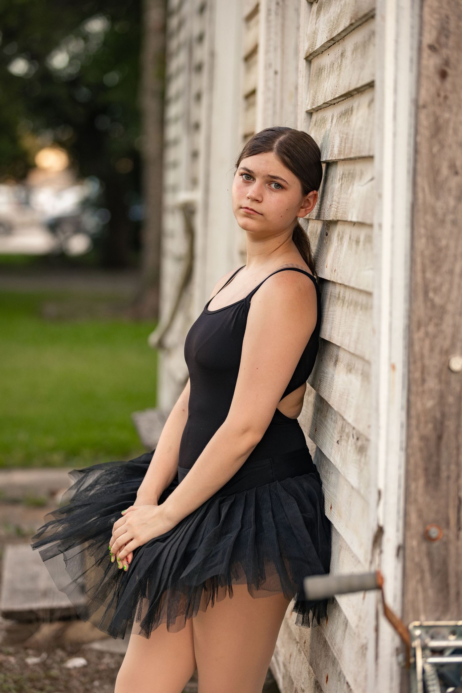 Young female ballet dancer in black tutu leaning against weathered wooden building wall outdoors.