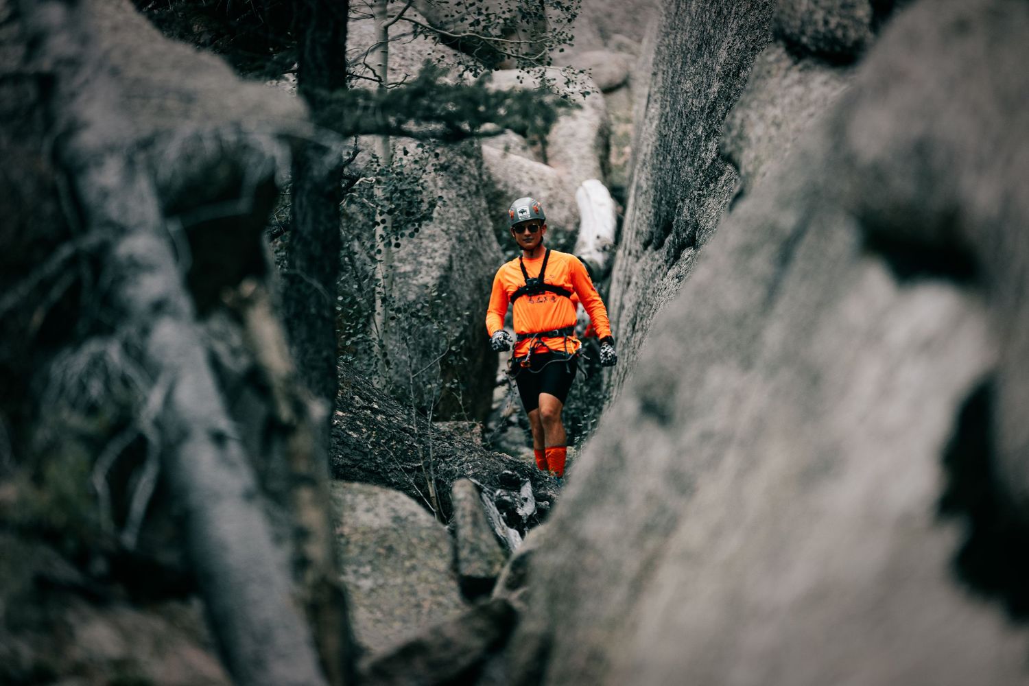 A rock climber in an orange jacket and helmet navigates through a narrow rocky crevice in a rugged mountain landscape.