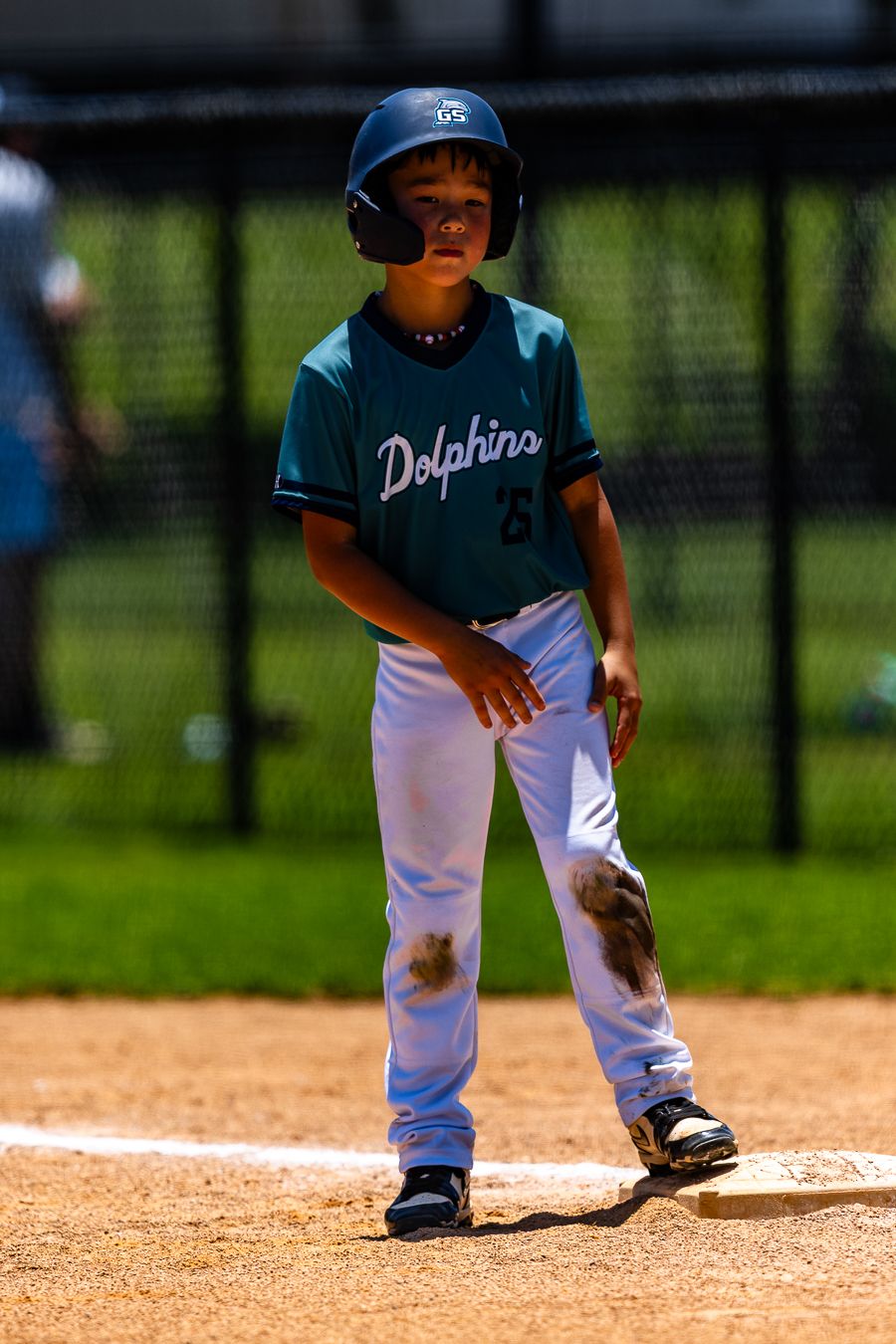 Young baseball player in teal Dolphins uniform standing on base with dirt-stained pants.