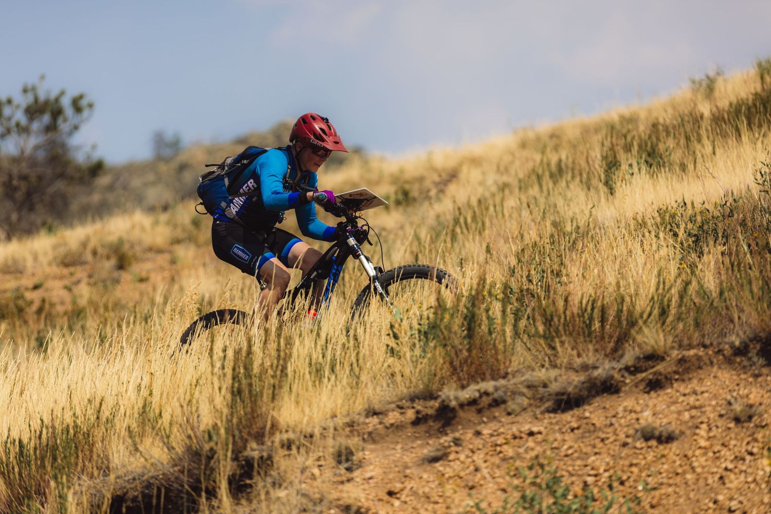 Mountain biker in red helmet riding through golden dry grass hillside trail on sunny day.