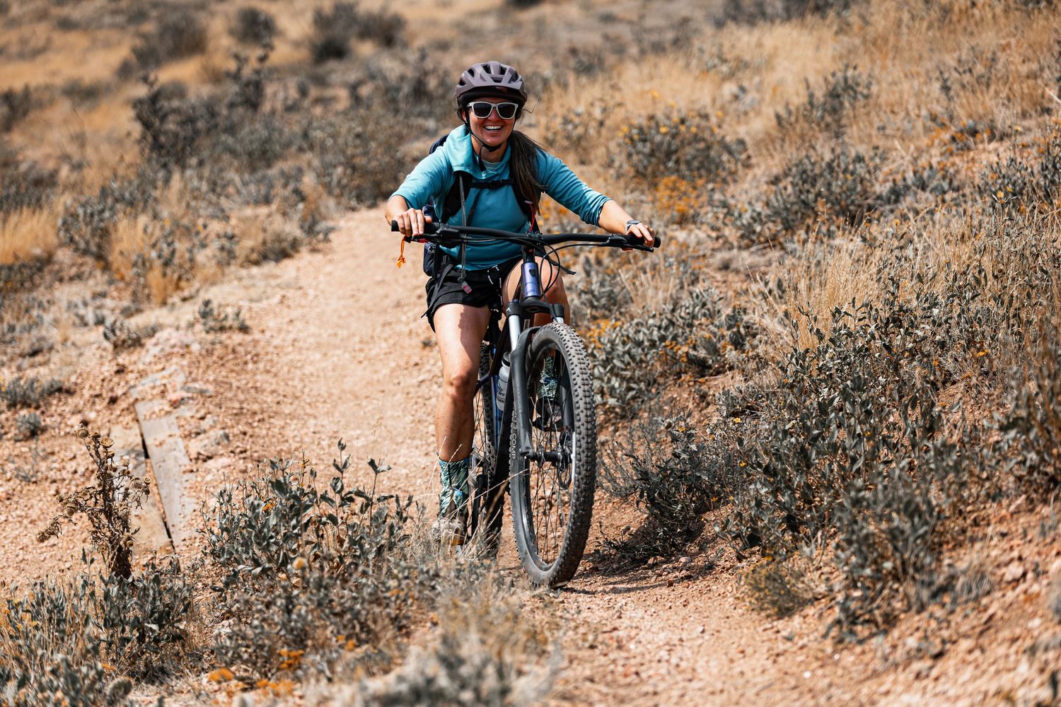 Woman mountain biking on dusty desert trail wearing teal jacket and white helmet outdoors.