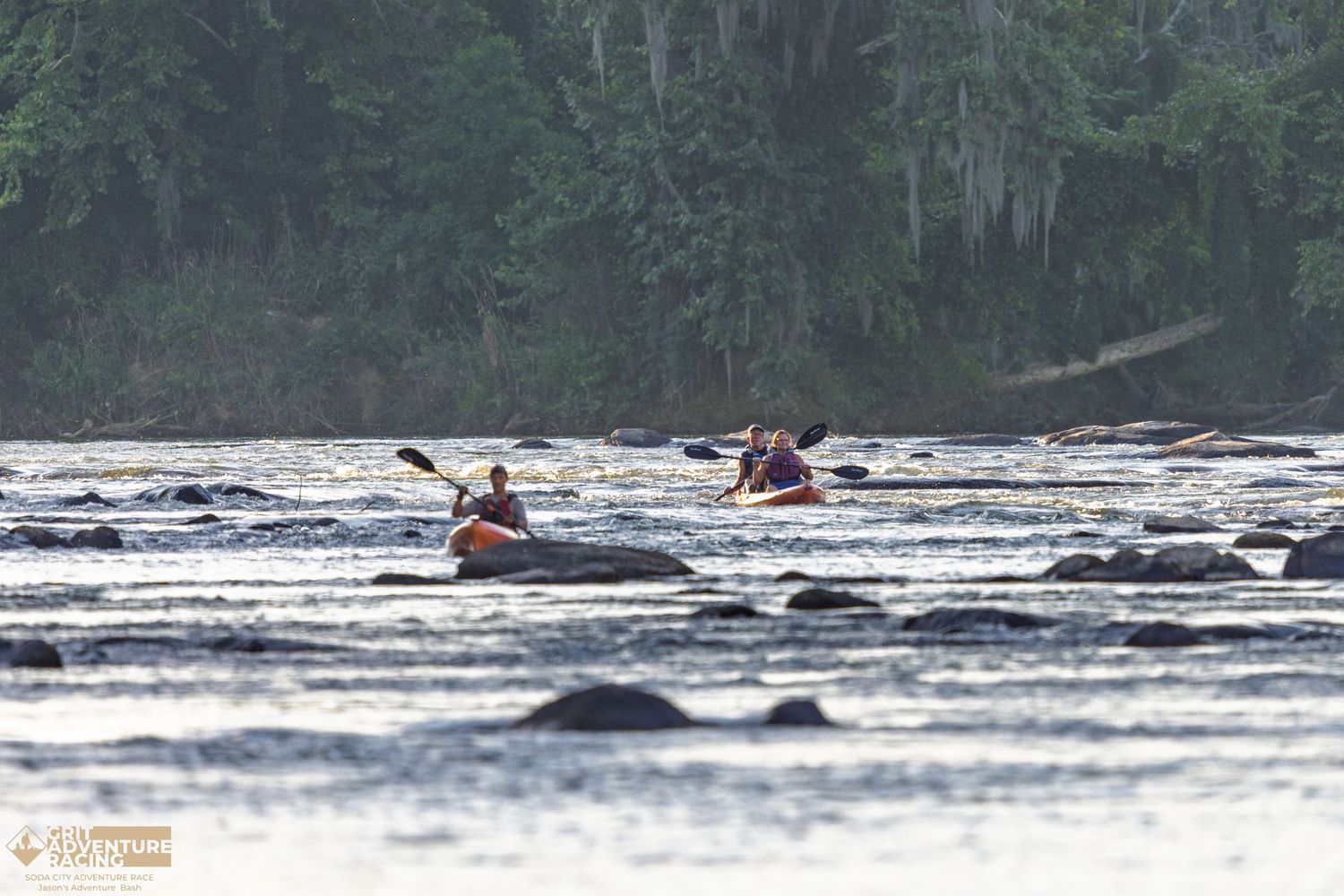 Two kayakers paddling through rocky river rapids surrounded by lush green forest trees.