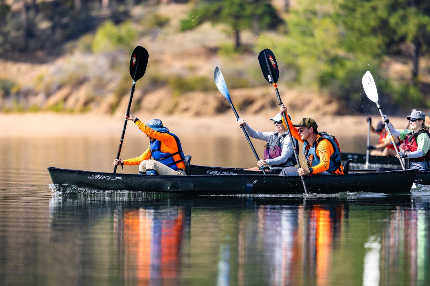 Group of three people kayaking in canoes on a calm lake with autumn hillside in background.