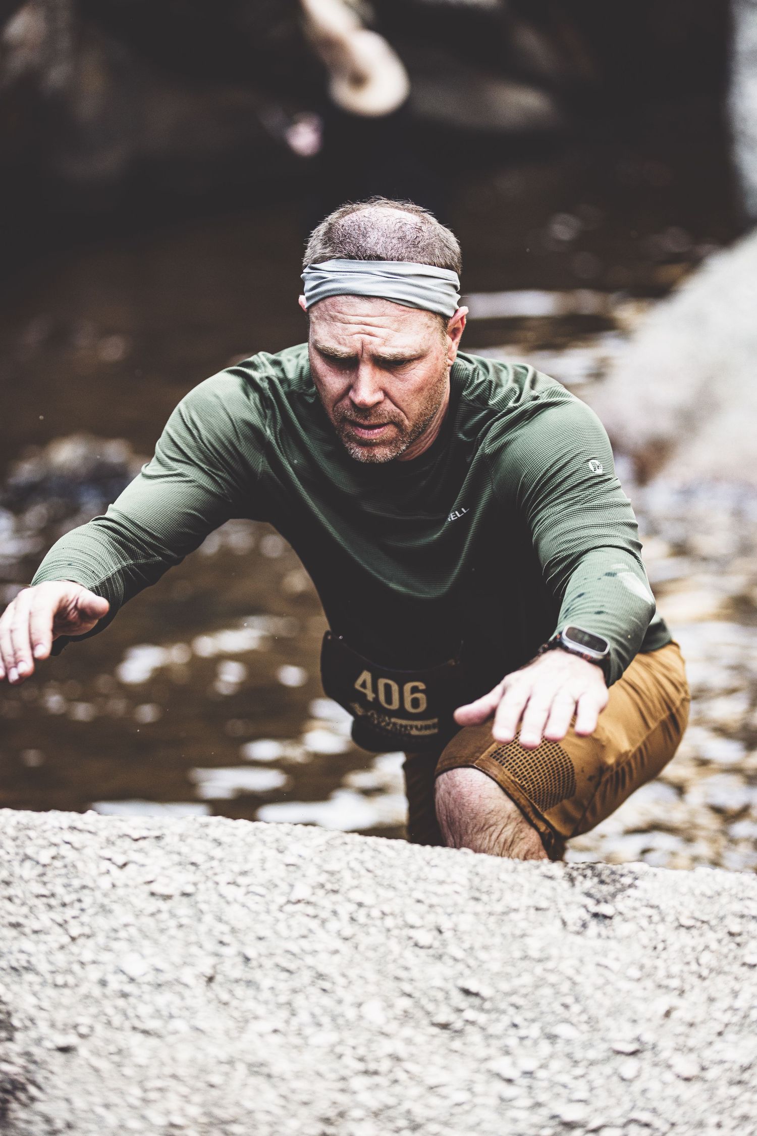 Determined male obstacle course racer number 406 climbing over wet rocks in a stream.
