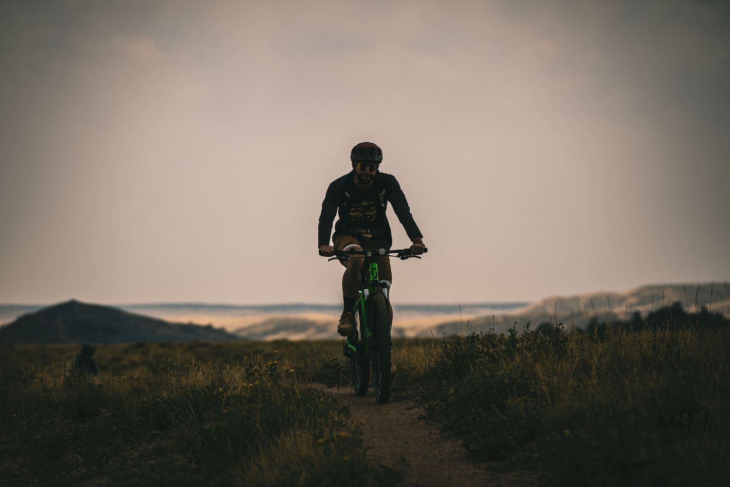 Silhouette of mountain biker pausing on hilltop trail at dusk with scenic landscape behind.