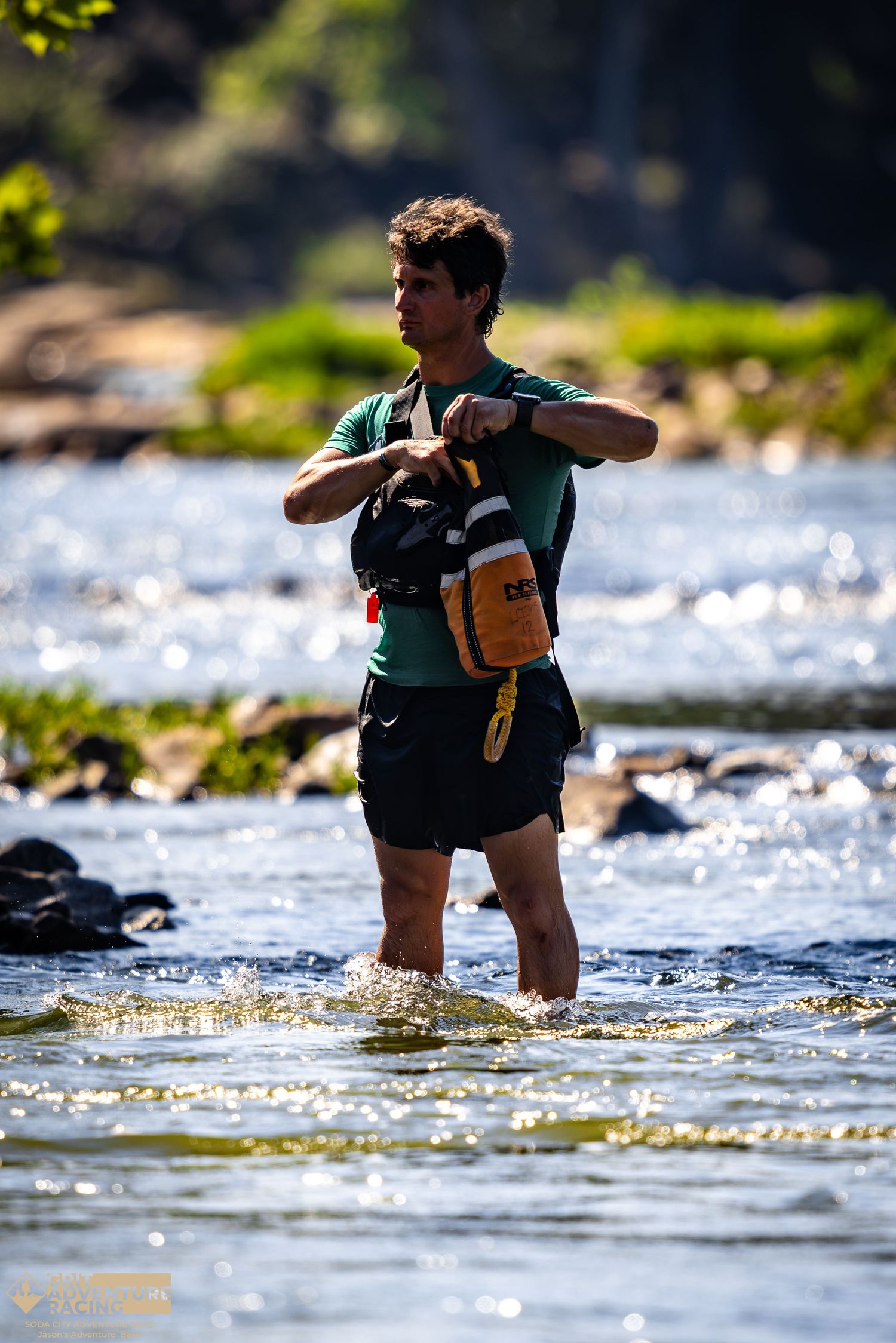 Athletic young man wading through a sunlit rocky river while adjusting his orange and black backpack.