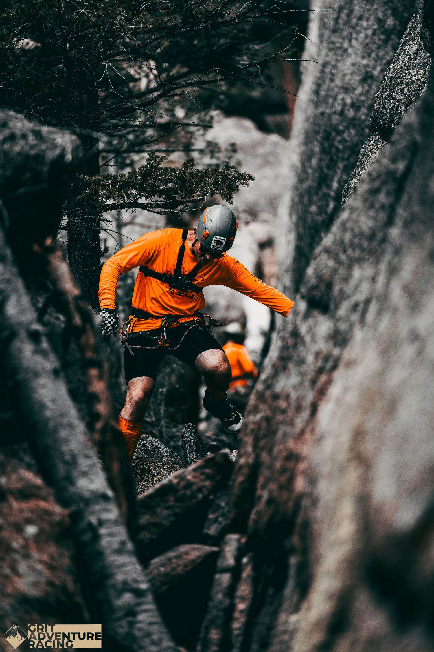 Adventure racer in orange shirt navigates rocky terrain through narrow tree-lined mountain passage.