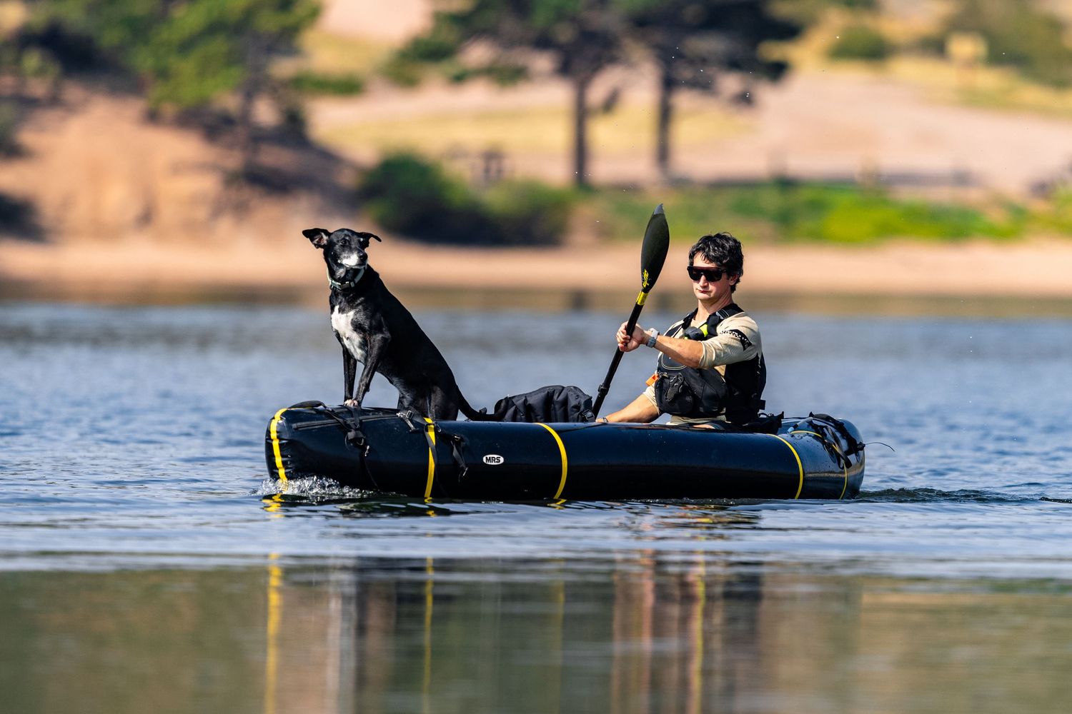 Man paddling inflatable kayak on calm lake with his black dog sitting at the bow.