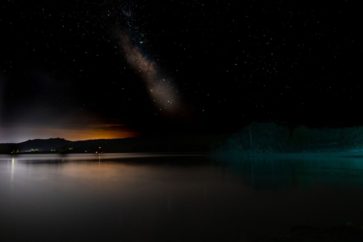 Stunning night sky with Milky Way galaxy over calm lake with glowing teal bioluminescence.