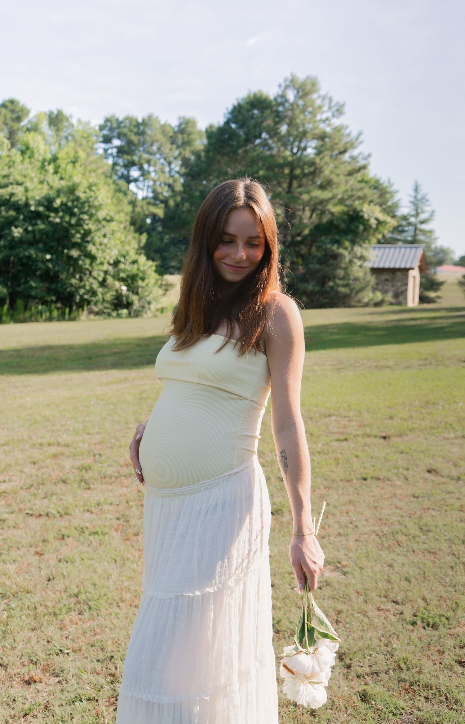 Pregnant woman in white dress holding flowers, smiling outdoors in a sunny green field.