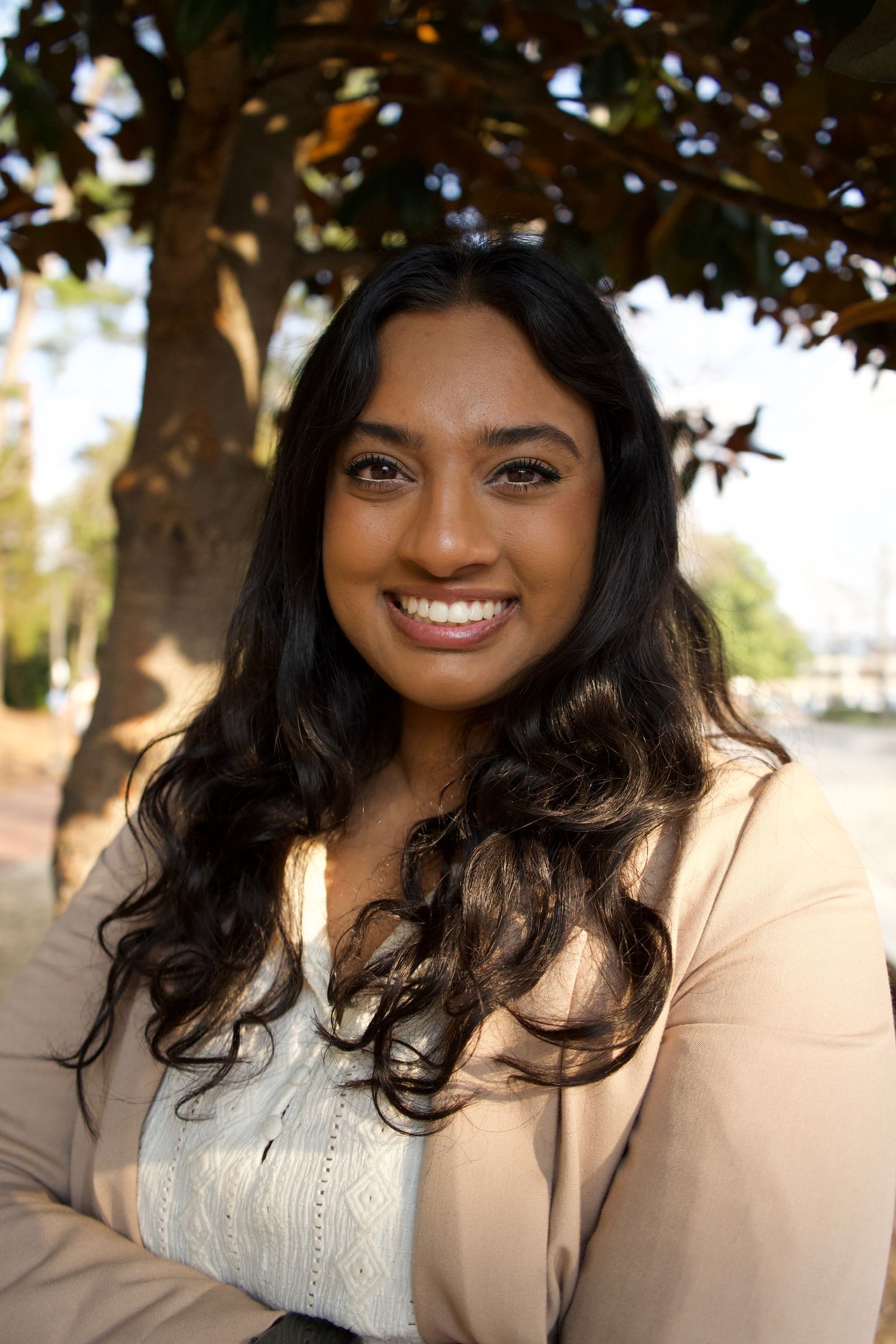 Smiling young woman in beige blazer with long dark wavy hair poses for portrait under tree outdoors.