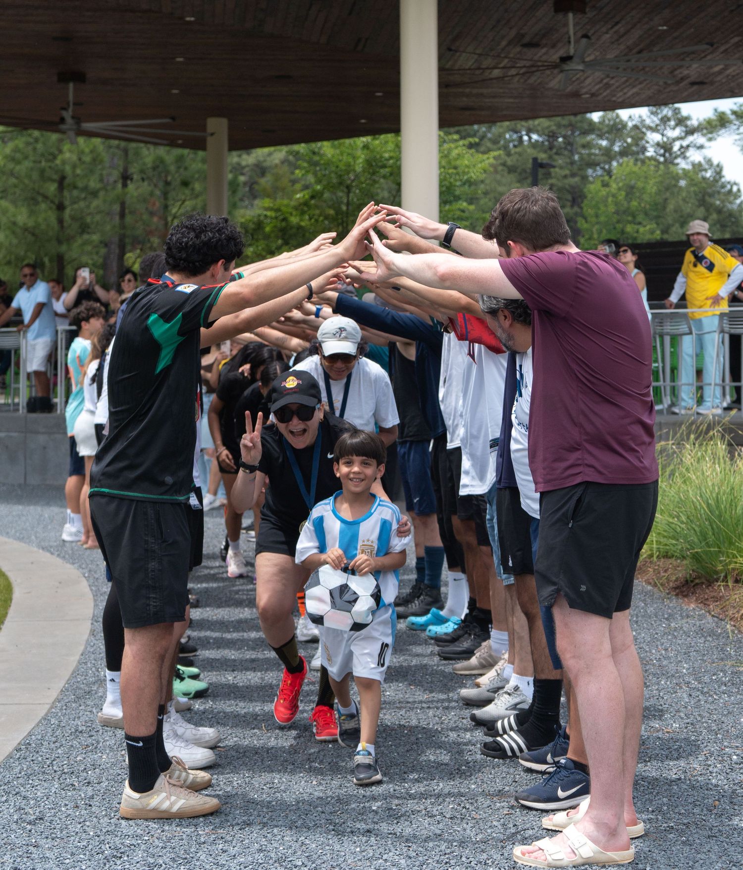Children and adults form a celebratory tunnel arch at an outdoor soccer event near a pavilion.