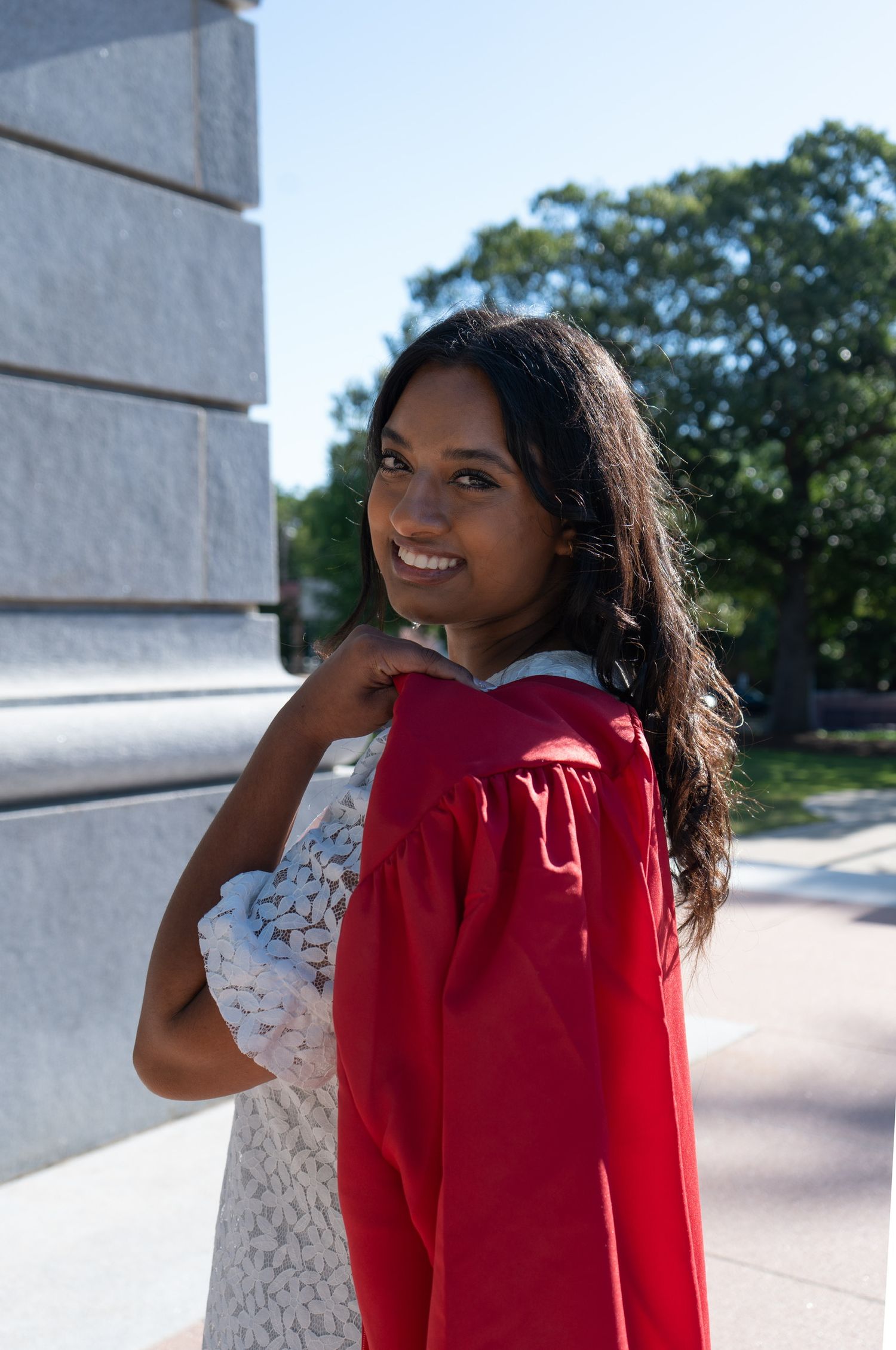 Smiling young woman in red graduation gown poses outdoors near stone column with trees in background.
