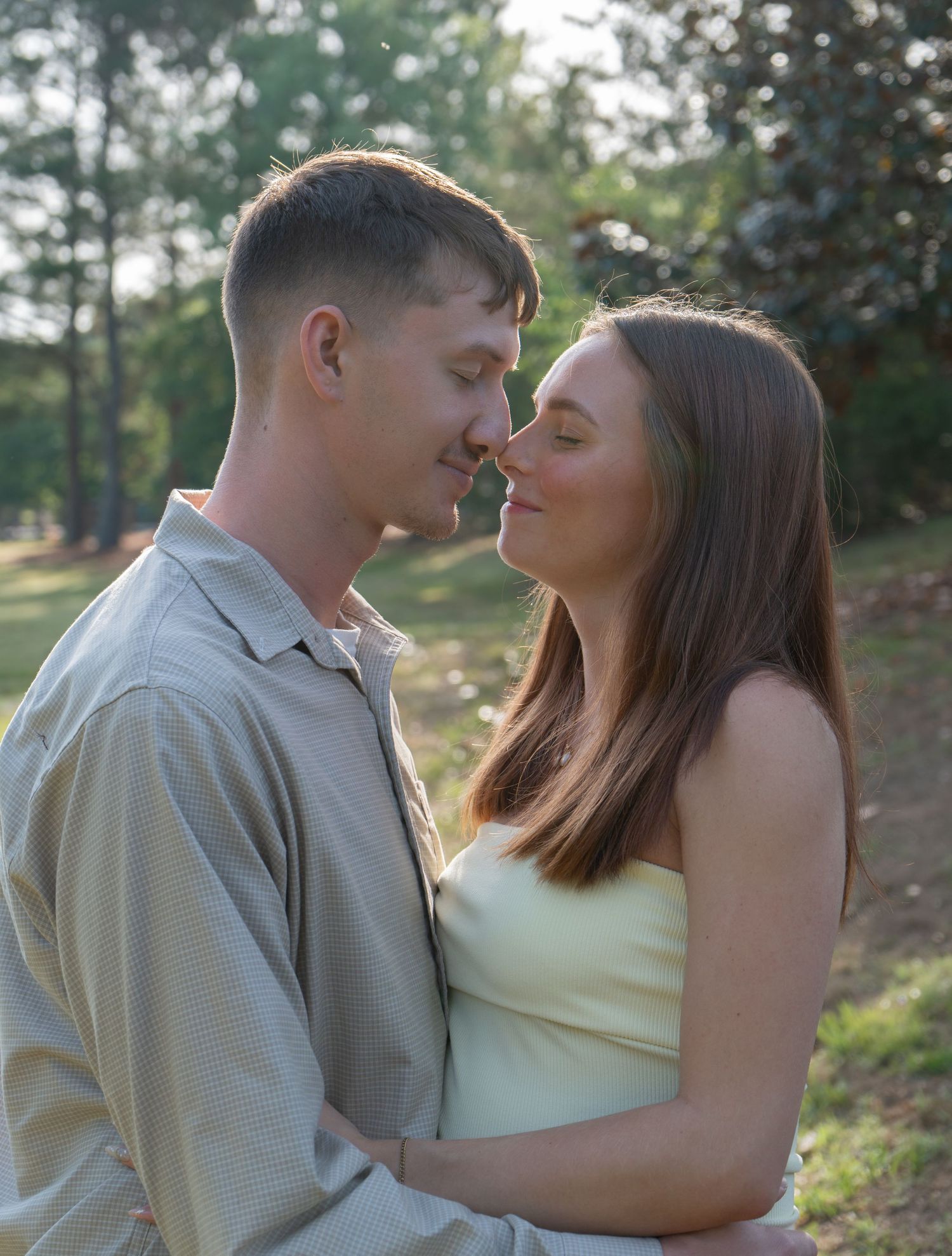 A young couple shares a romantic moment outdoors, nearly touching noses, surrounded by lush green trees.