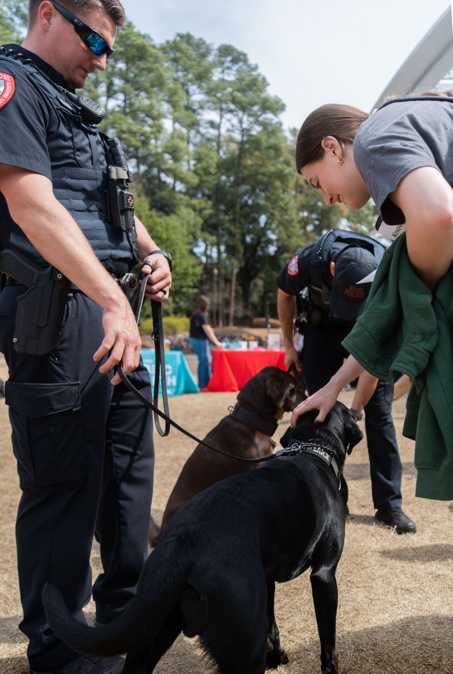Police officer and student interact with a black K9 dog on a leash at an outdoor campus event.