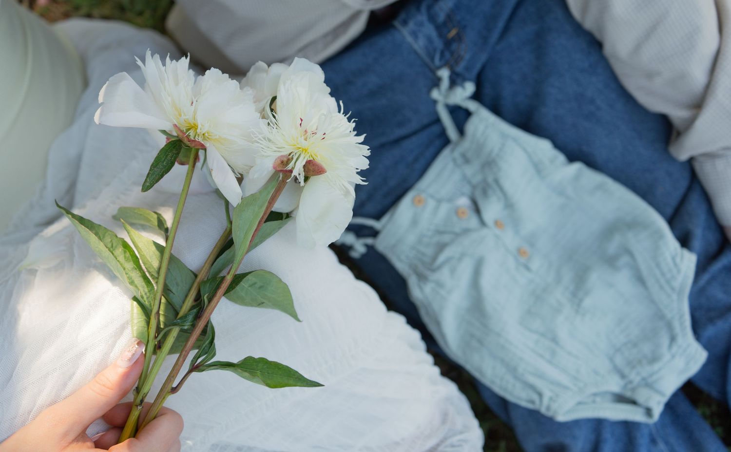 Hand holding white peony flowers next to folded blue denim and light blue baby bloomers outdoors.