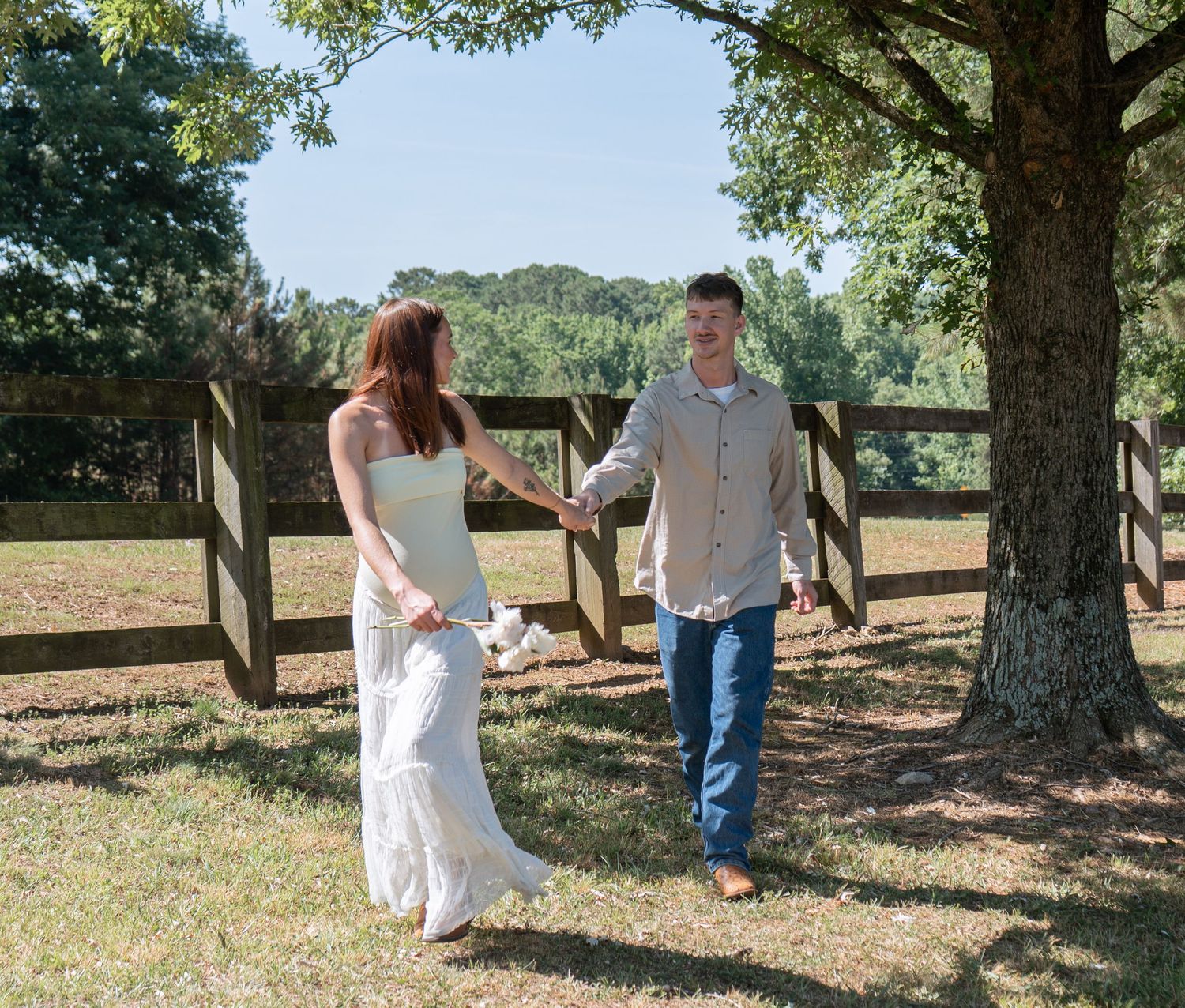 Couple holding hands in a sunny farm field near a wooden fence and large tree on their wedding day.