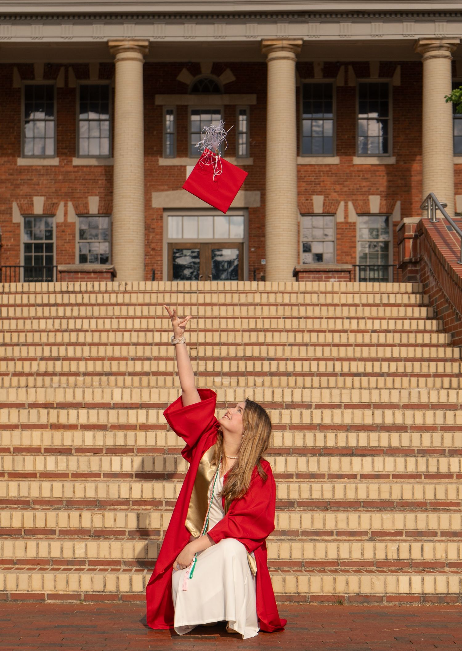 Graduate in red cap and gown tossing mortarboard on brick steps of a university building.