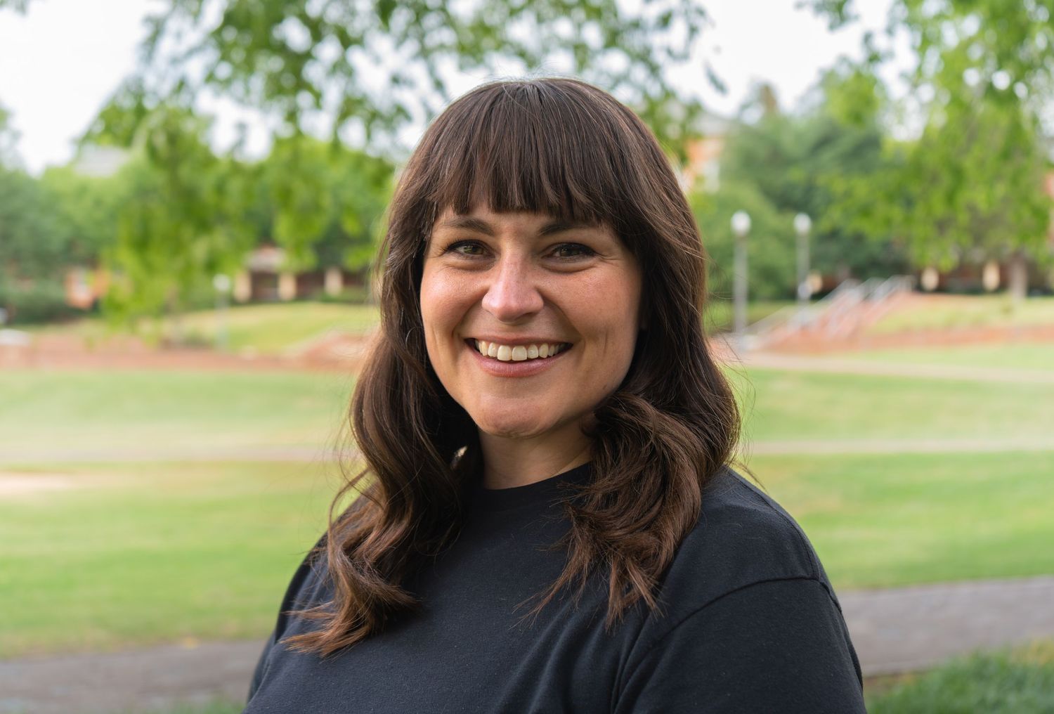 Smiling woman with dark hair and bangs wearing black shirt standing outdoors in green park setting.