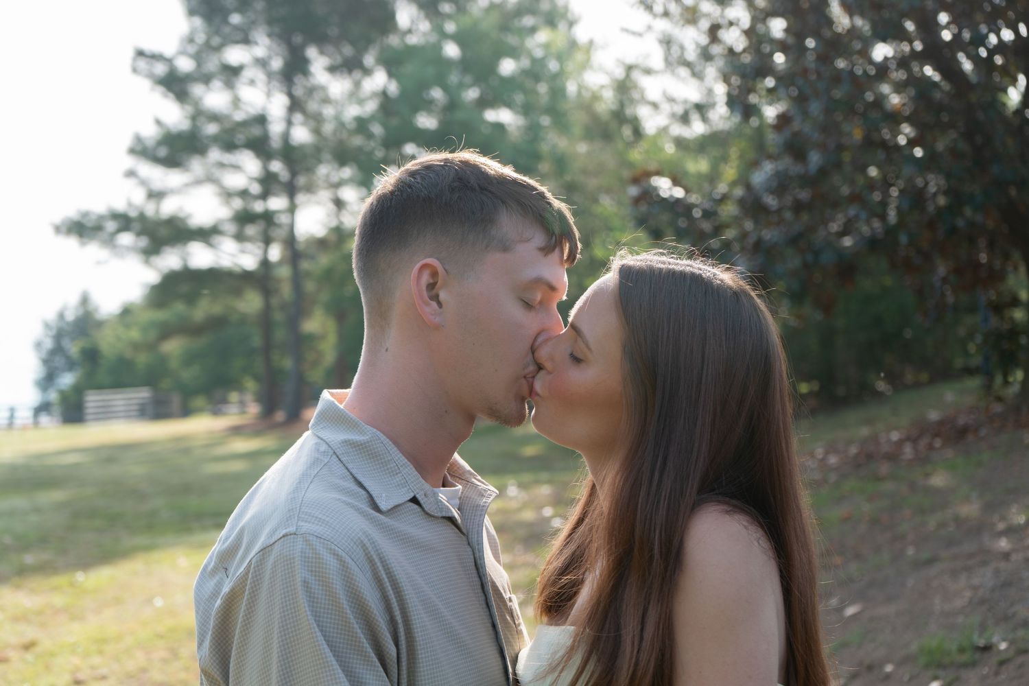 A young couple sharing a romantic kiss outdoors in a sunny park surrounded by green trees.