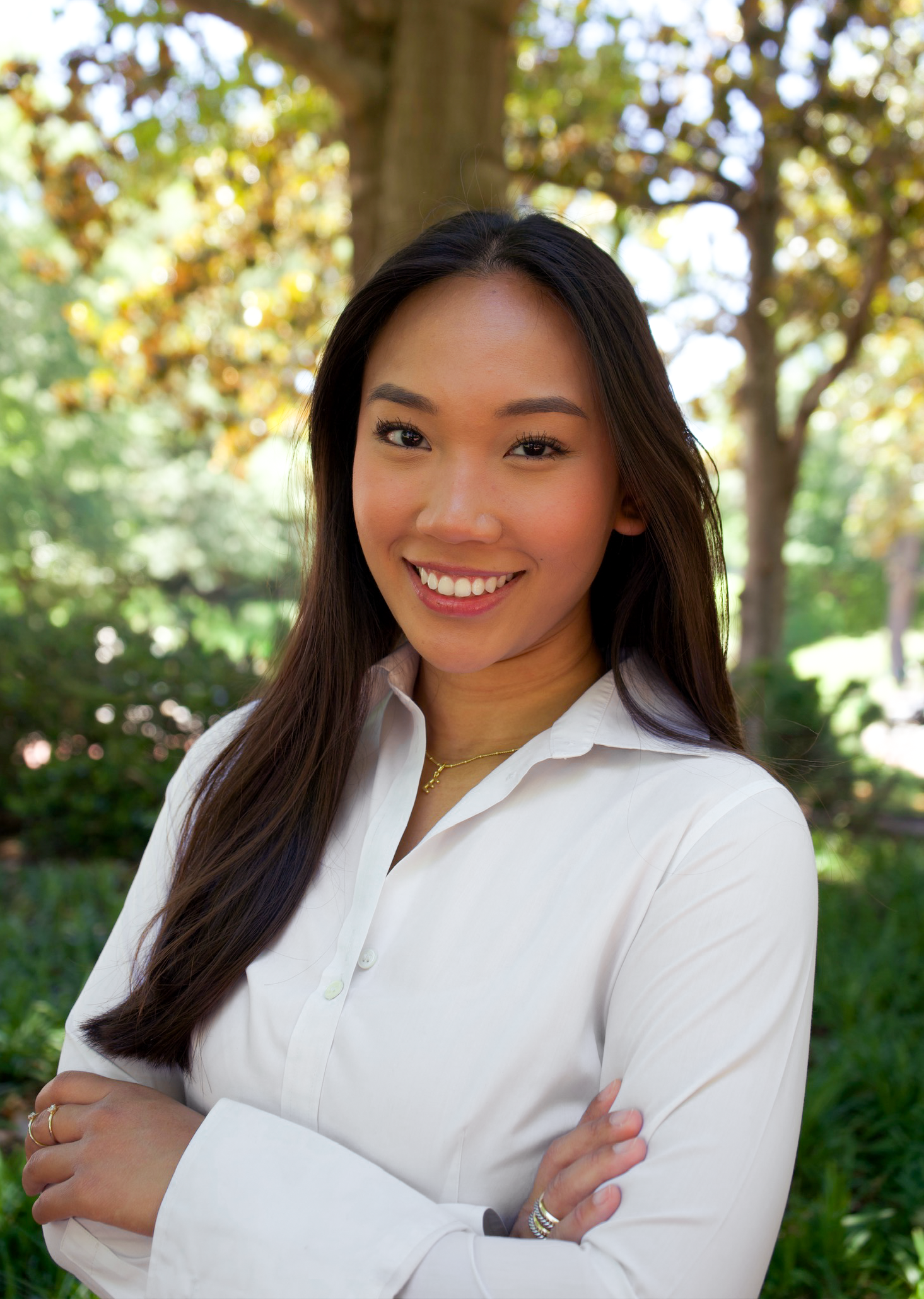 Young woman with long dark hair smiling outdoors in white shirt with arms crossed among green trees.