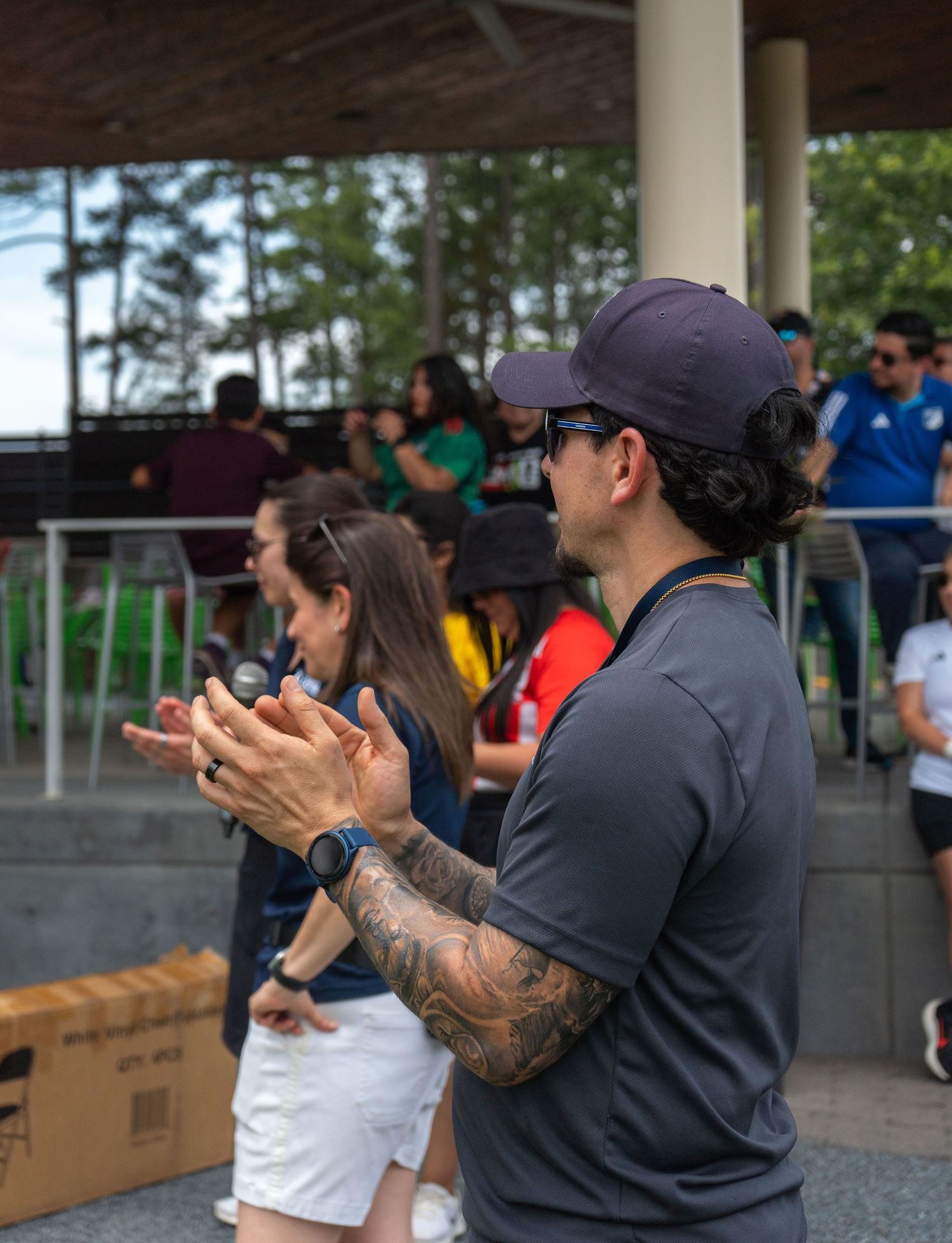 Tattooed man in gray shirt and cap clapping at an outdoor sports event near a pavilion captured by Triangle photographer.