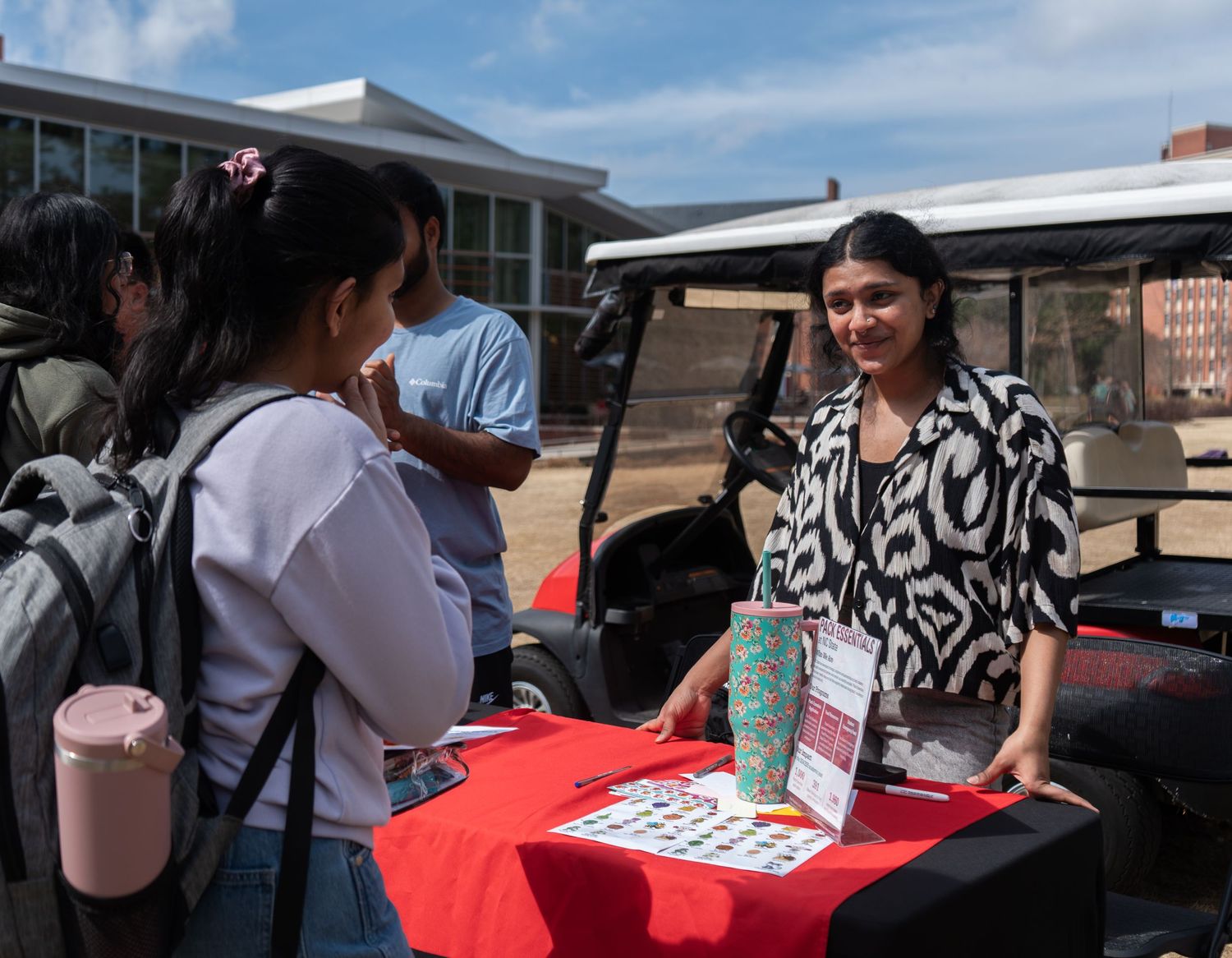 Student representative in black and white top speaks with visitors at a red NC State table.