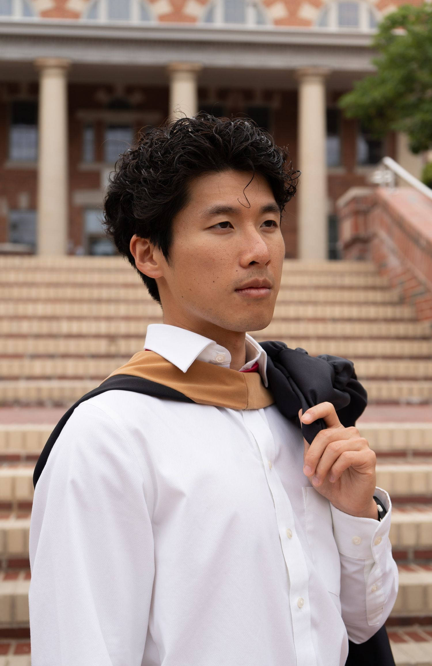 Young man in white shirt carries graduation gown over shoulder on university steps with columns.