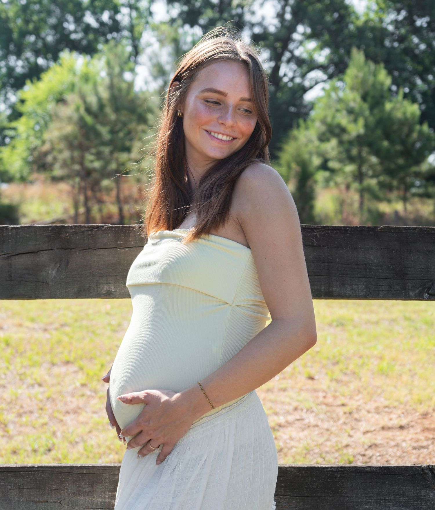 Smiling pregnant woman in a yellow tube top dress cradling her baby bump outdoors near a wooden fence.
