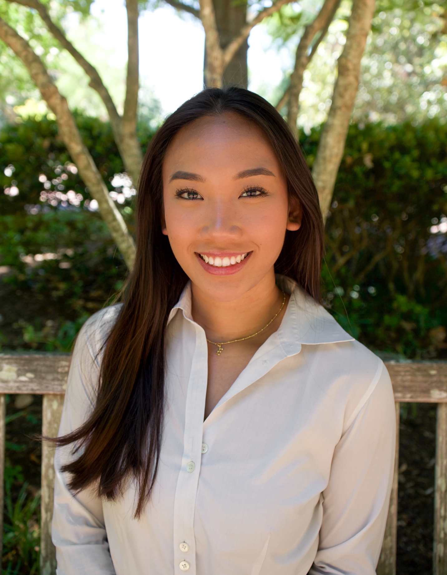 Young woman with long dark hair smiling warmly in a light shirt outdoors with trees in background.