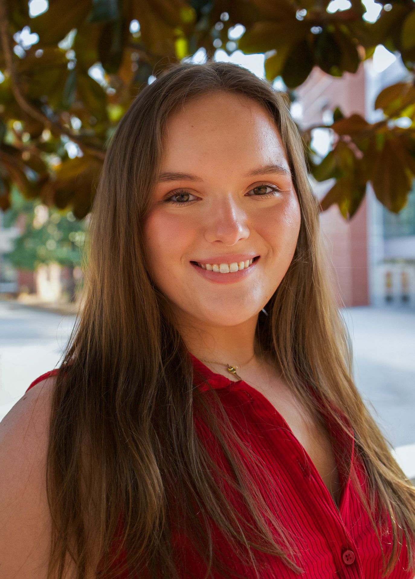 A smiling young woman with long brown hair poses outdoors in a red top beneath green foliage.