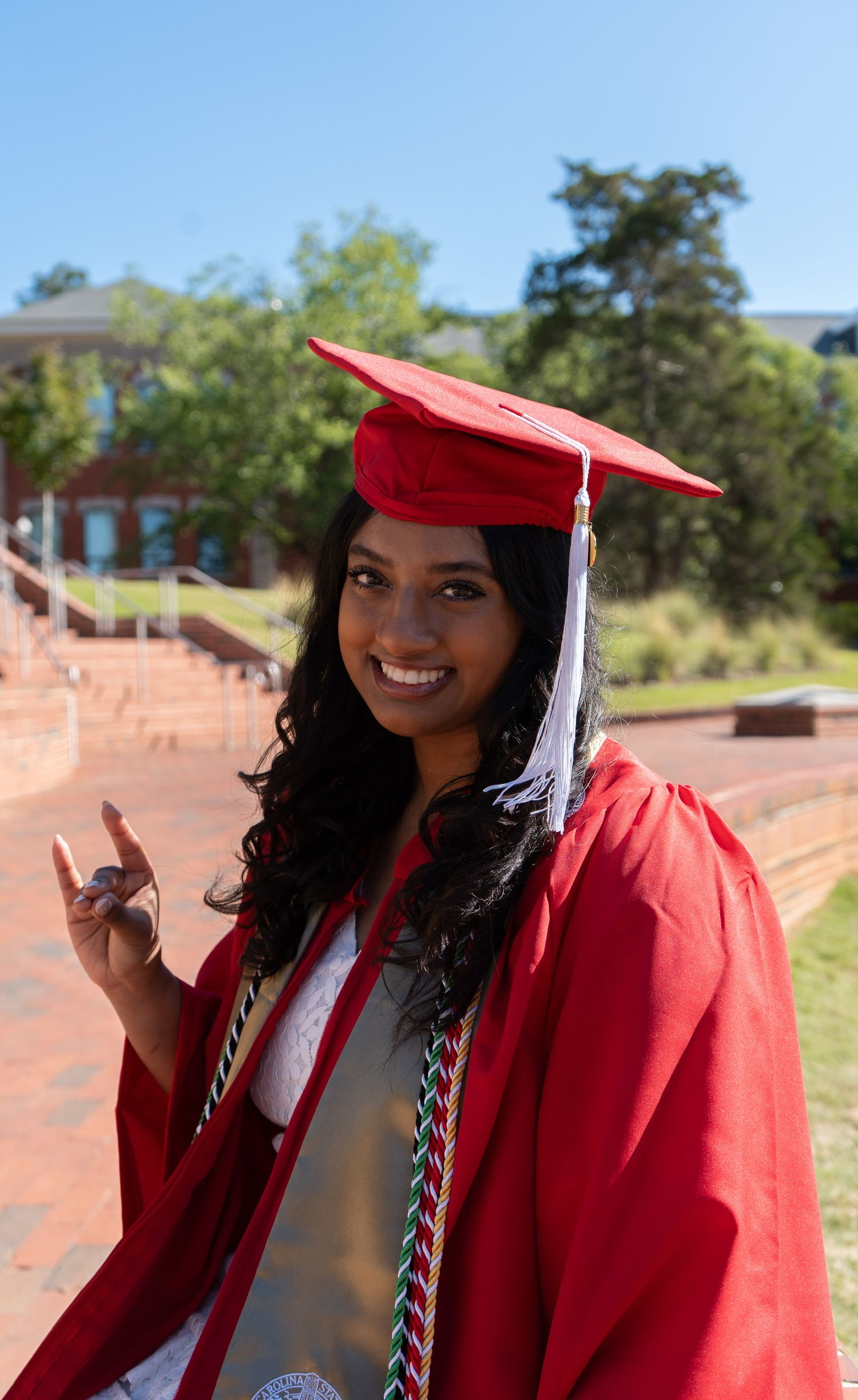 Smiling NC State graduate in red cap and gown with honor cords poses outdoors on brick plaza on sunny day.